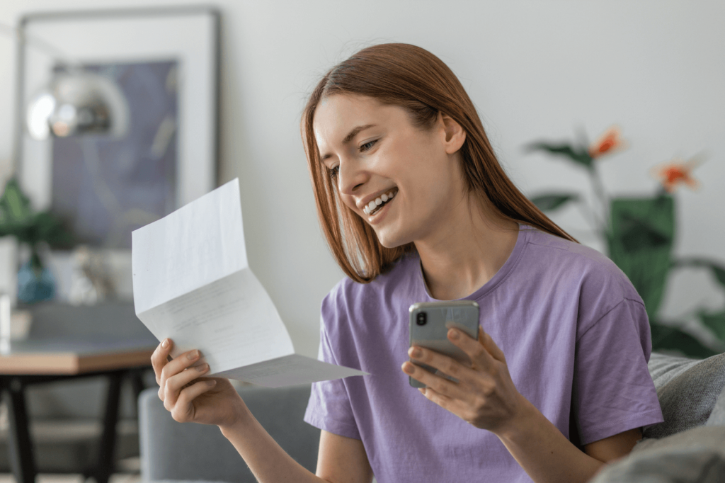A woman smiling while managing her bills using a smartphone.