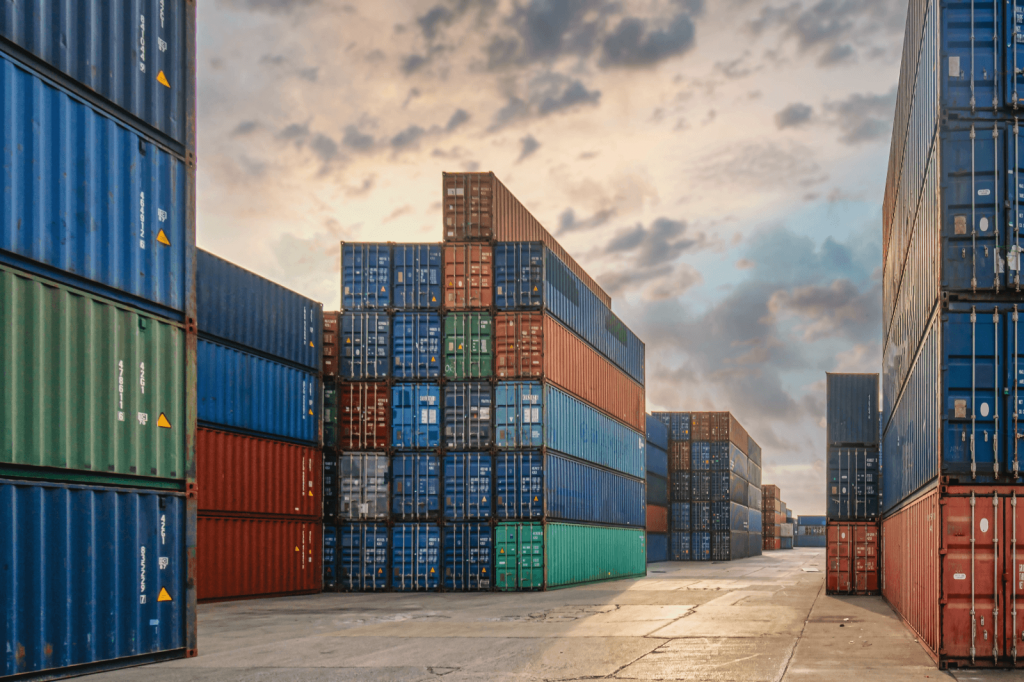 Stacked shipping containers at a port under a cloudy sky