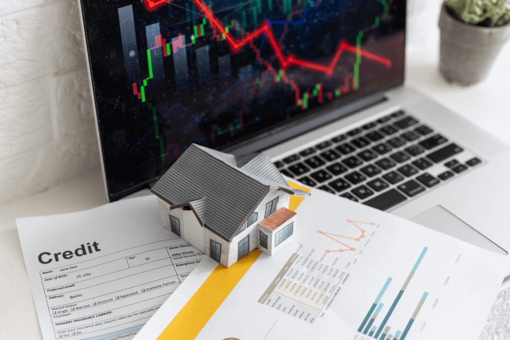 A house on documents with a downward graph on a laptop screen.