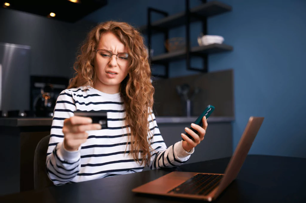 Confused woman looking at her credit card while holding a phone